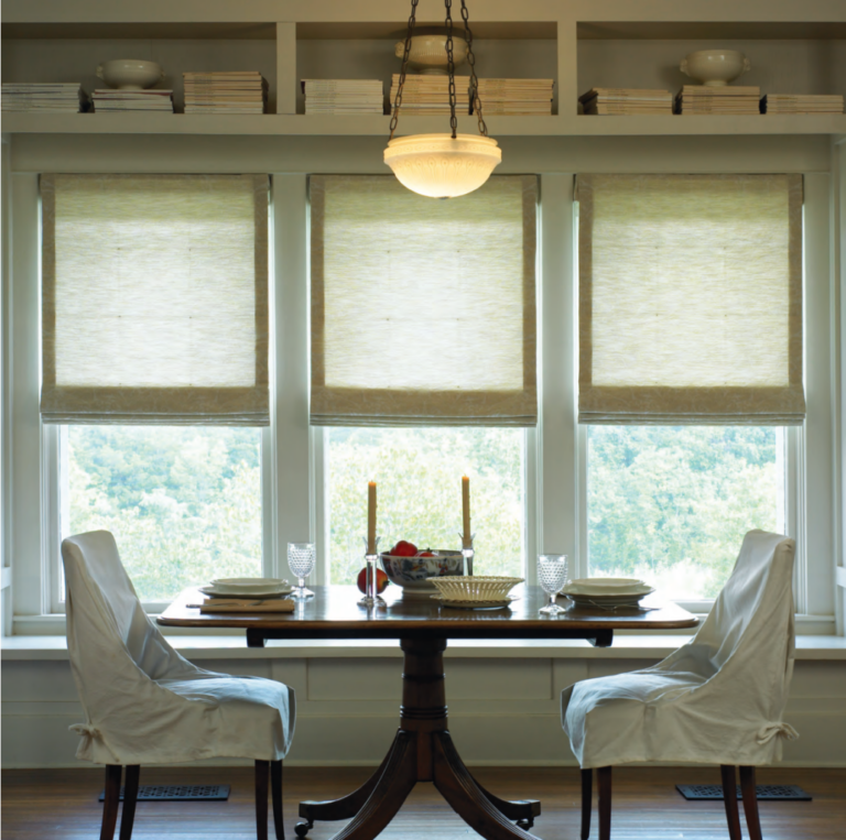 Roman shades in a woven fabric lowered across three dining room windows beneath a pendant light