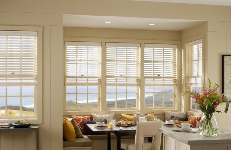 Cream wood blinds tilted to filter light across a bay-window breakfast nook with an ocean view