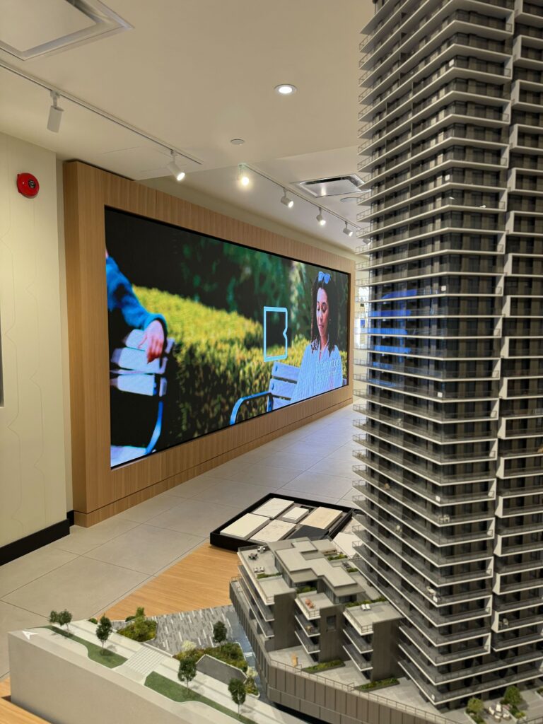 Wide-angle shot of Pure Image’s custom video wall in a Vancouver sales gallery, displaying vivid digital content alongside a detailed architectural scale model.