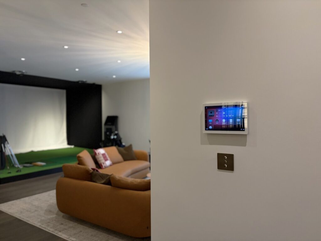 Wall-mounted Control4 touchscreen and Lutron Alisse keypad controlling a smart home lounge with a golf simulator, installed by Pure Image.