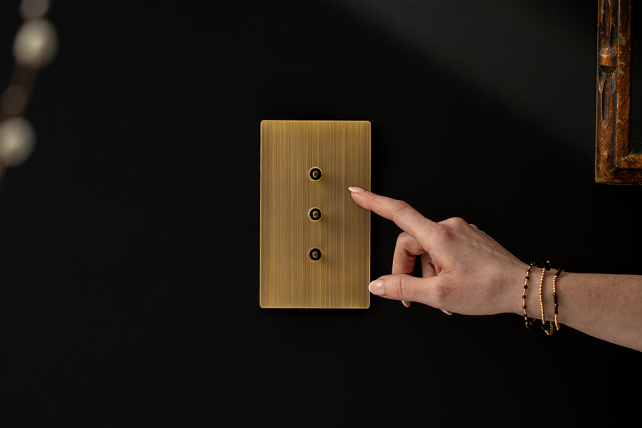 A hand pressing the middle toggle of a three-button Lutron Aviena keypad in Aged Brass on a matte black wall